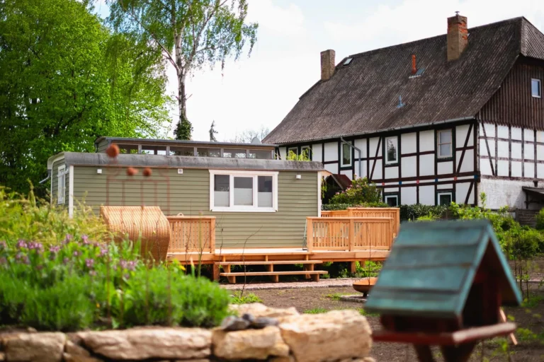 Tiny House Mimi auf dem Ilsehof mit Holzveranda und Strandkorb, eingebettet in Naturgarten mit Blick auf das historische Fachwerkhaus im Hintergrund