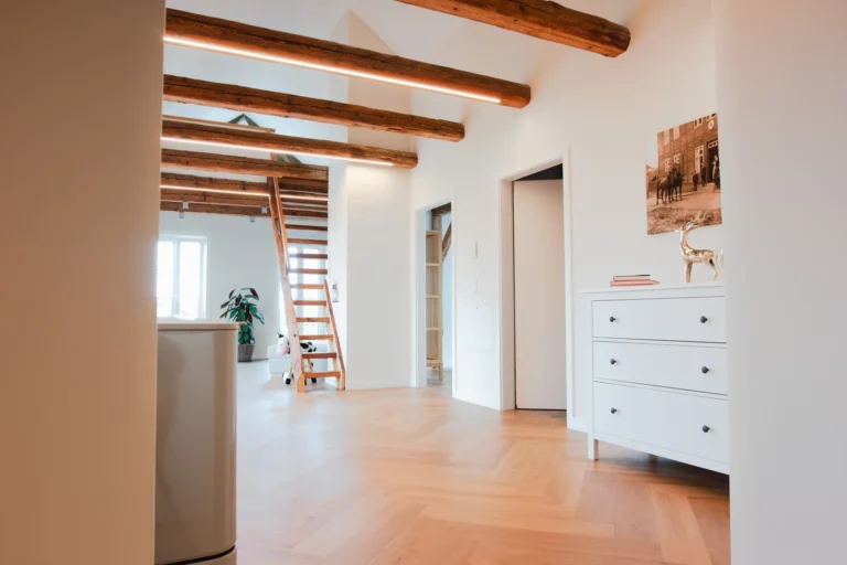 Blick in die Ferienwohnung Berta mit Sicht auf den offenen Wohnbereich, Holztreppe zum offenen Schlafbereich, Holzbalken-Decke und weißer Kommode im Eingangsbereich.
