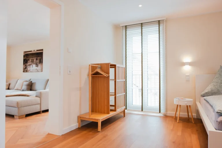 Schlafzimmer der Ferienwohnung Ruth mit Doppelbett, Kleiderschrank aus Holz und Blick ins Wohnzimmer mit grauer Couch.