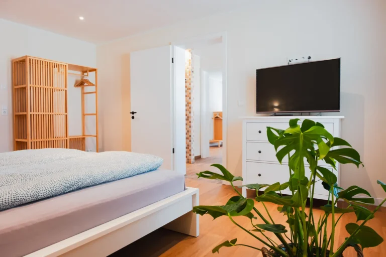 Schlafzimmer mit Doppelbett, Holzkleiderschrank und Blick auf einen Fernseher auf weißer Kommode.